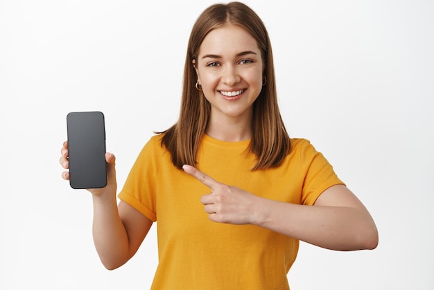 Retrato de mujer joven apuntando a la venta en la pantalla del teléfono inteligente que muestra la interfaz de la aplicación de visualización del teléfono móvil sonriendo satisfecha de pie en camiseta amarilla sobre fondo blanco