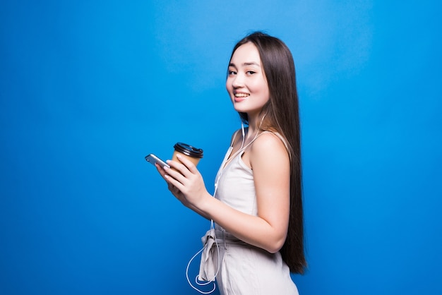 Retrato de mujer joven asiática sonrisa permanente, utilizando el teléfono móvil, sosteniendo la taza de papel de café, mirando smartphone en la pared azul