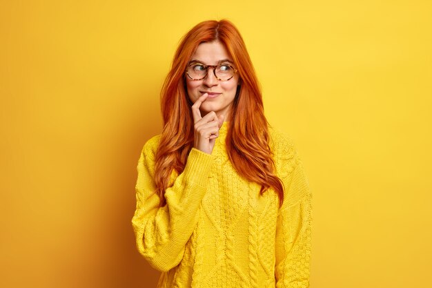 El retrato de una mujer joven de aspecto agradable y complacida mantiene el dedo índice cerca de los labios concentrado a un lado, tiene una expresión pensativa, el cabello rojo natural usa un suéter casual.