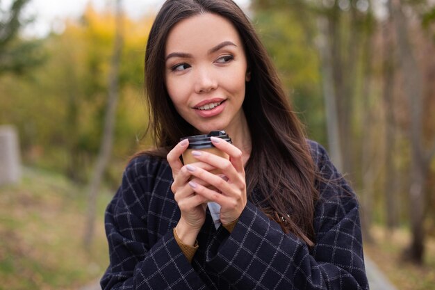 Retrato de una mujer joven y atractiva con abrigo y café para ir a hurtadillas mirando hacia otro lado en el parque de otoño