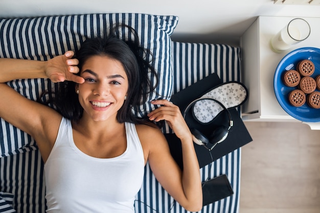 Retrato de mujer joven y atractiva acostada en la cama sonriendo, vista desde arriba, despertarse por la mañana, perezoso, tiempo libre, domingo, relajante, desayuno, teléfono, auriculares, libro, antifaz para dormir