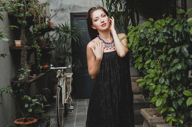 Retrato de mujer joven y atractiva en elegante vestido negro posando en villa tropical, estilo de verano sexy y elegante, accesorios de collar de moda, sonriendo, joyas, piel pura y natural de la cara