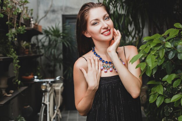 Retrato de mujer joven y atractiva en elegante vestido negro posando en villa tropical, estilo de verano sexy y elegante, accesorios de collar de moda, sonriendo, joyas, piel pura y natural de la cara