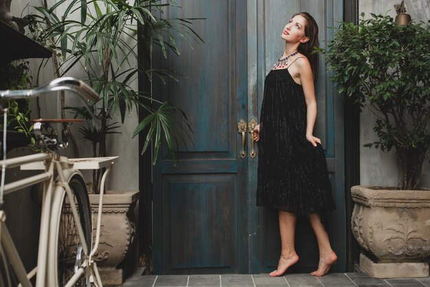 Retrato de mujer joven y atractiva en elegante vestido negro posando en villa tropical, estilo de verano sexy y elegante, accesorios de collar de moda, sonriente, estado de ánimo romántico, lujo