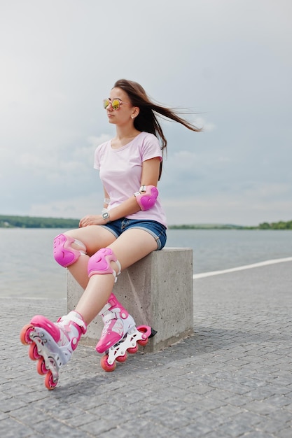 Retrato de una mujer joven y atractiva con pantalones cortos, camiseta, gafas de sol y patines sentados en el banco de hormigón en la pista de patinaje al aire libre