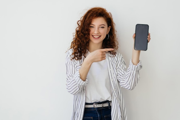 Retrato de una mujer joven y atractiva con el pelo rojo largo y rizado que se encuentra aislada sobre un fondo blanco que muestra un teléfono móvil con una pantalla en blanco
