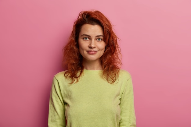 Retrato de mujer joven atractiva tiene cabello rojo natural, mira directamente a la cámara con una sonrisa suave