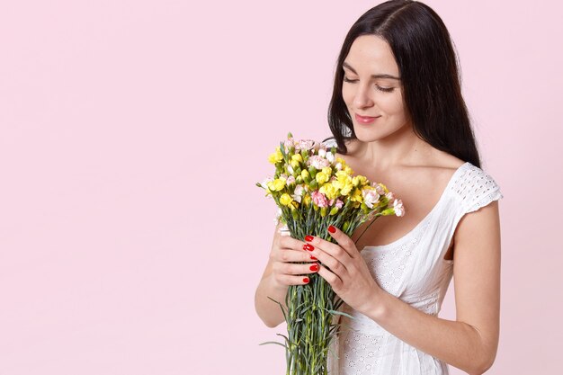 Retrato de mujer joven atractiva tierna con largo cabello negro en vestido blanco de verano con ramo, oliendo flores