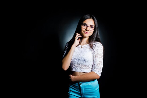 Retrato de una mujer joven atractiva en top blanco y pantalones azules posando con sus gafas en la oscuridad