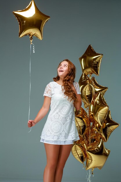 Retrato de una mujer joven y atractiva con un vestido blanco, sosteniendo un montón de globos dorados, sobre un fondo claro