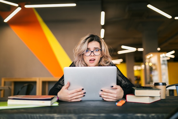 Retrato de mujer joven bonita con expresión de la cara calcetín, sentado en la mesa trabajando en la computadora portátil