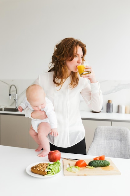 Retrato de mujer joven y bonita de pie y sosteniendo a su pequeño bebé mientras bebe jugo y cocina en la cocina