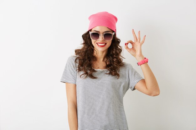 Retrato de mujer joven y bonita que muestra bien firmar, con sombrero rosa, gafas de sol, sonriendo, aislado