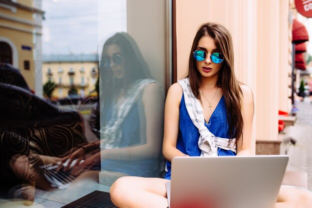 Retrato de mujer joven y bonita sentada durante el día de verano mientras usa una computadora portátil y un auricular inalámbrico para videollamadas