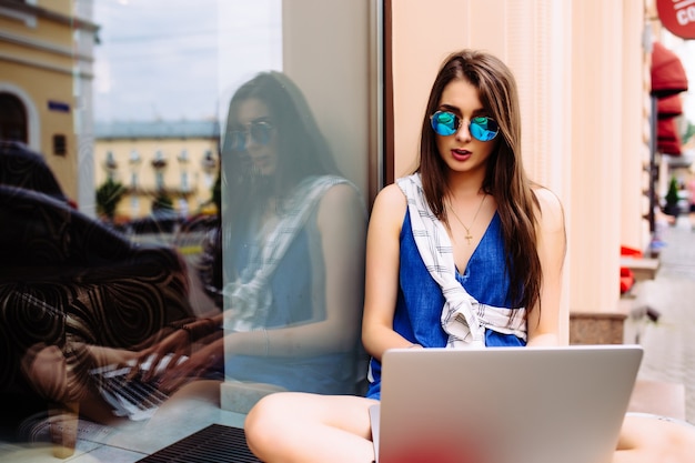 Retrato de mujer joven y bonita sentada durante el día de verano mientras usa una computadora portátil y un auricular inalámbrico para videollamadas