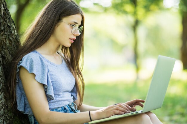 Retrato de mujer joven y bonita sentada sobre la hierba verde en el parque con las piernas cruzadas durante el día de verano mientras usa la computadora portátil
