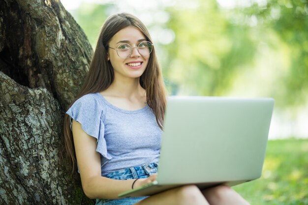 Retrato de mujer joven y bonita sentada sobre la hierba verde en el parque con las piernas cruzadas durante el día de verano mientras usa la computadora portátil