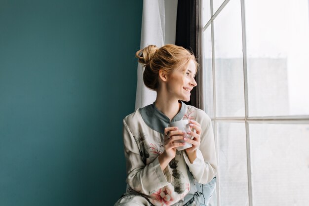 Retrato de mujer joven con cabello rubio tomando café o té junto a la ventana grande, sonriendo, disfrutando de la feliz mañana en casa. Pared turquesa. Vistiendo pijamas de seda con flores.