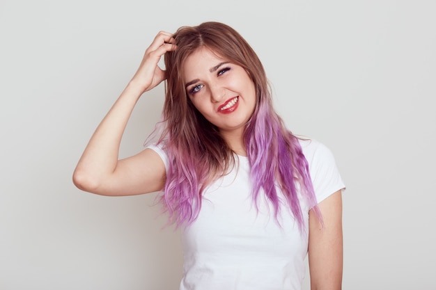 Retrato de mujer joven en camiseta blanca de estilo casual rascándose el pelo de la caspa y la irritación, que sufre de piojos, con el ceño fruncido, aislado sobre fondo gris.