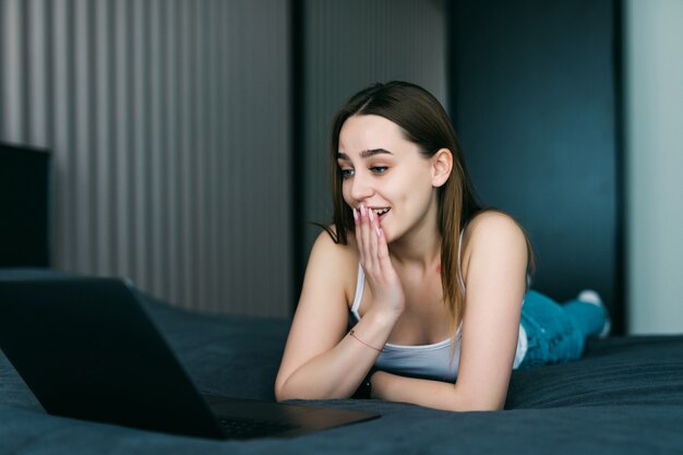 Retrato de una mujer joven casual sorprendida usando la computadora portátil en la cama