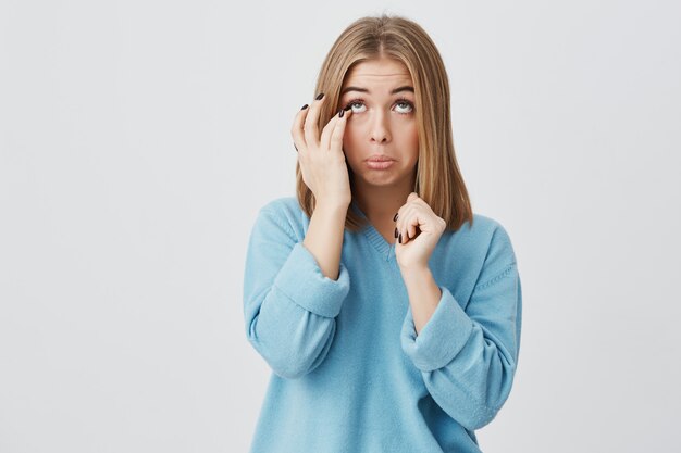 Retrato de mujer joven caucásica con cara ovalada, ojos oscuros y cabello lacio y rubio con un suéter casual azul, tocando su rostro, tratando de sacar las pestañas de sus ojos, mirando hacia arriba.