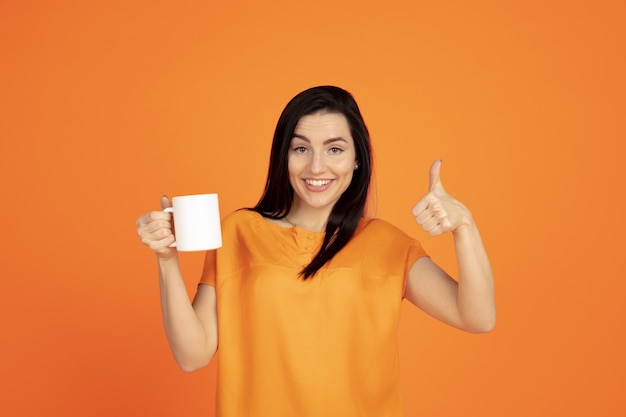 Retrato de mujer joven caucásica sobre fondo naranja de estudio. Modelo morena mujer hermosa en camisa. Concepto de emociones humanas, expresión facial, ventas, publicidad. Copyspace. Beber café o té.