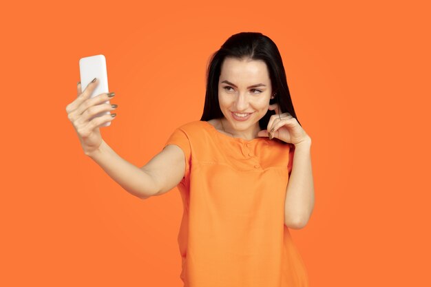 Retrato de mujer joven caucásica sobre fondo naranja de estudio. Modelo morena mujer hermosa en camisa. Concepto de emociones humanas, expresión facial, ventas, publicidad. Copyspace. Haciendo selfie, gana en apuesta.