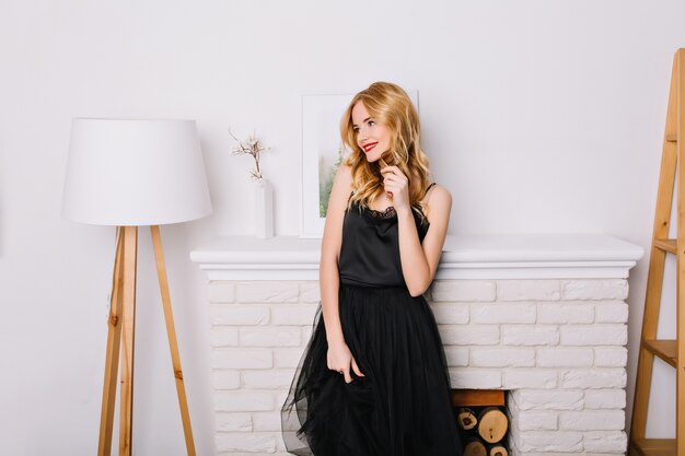 Retrato de mujer joven, chica rubia en una habitación luminosa con un interior blanco agradable y moderno, de pie junto a una chimenea falsa, mirando al lado. El uso de un elegante vestido negro.