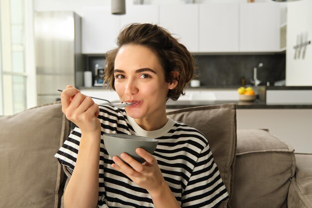 Retrato de una mujer joven comiendo una taza de granola de cereales con leche sentada en el sofá y teniendo su