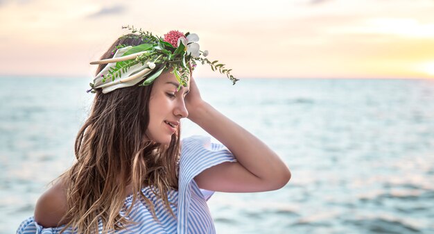 Retrato de una mujer joven con una composición de flores en la cabeza contra el fondo del océano al atardecer.