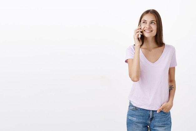 Retrato de mujer joven confiada feliz y despreocupada socisble con tatuaje en camiseta blanca sonriendo encantado mirando hacia arriba mientras sostiene el teléfono inteligente cerca de la oreja hablando por teléfono celular con un amigo