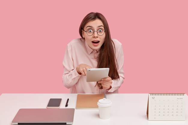 Retrato de mujer joven conmocionada hace trabajo remoto, sostiene el panel táctil, investiga el proyecto, mantiene la boca abierta