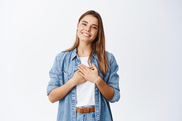 Retrato de mujer joven conmovida que se siente halagada, cogidos de la mano en el corazón y sonrientes dientes blancos, decir gracias, apreciar la ayuda, estar agradecido por un gesto agradable, blanco