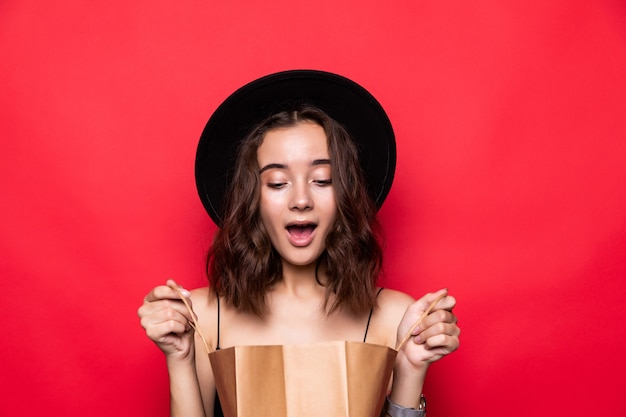 Retrato de una mujer joven curiosa con sombrero de verano mirando dentro de bolsas de compras aisladas sobre pared roja