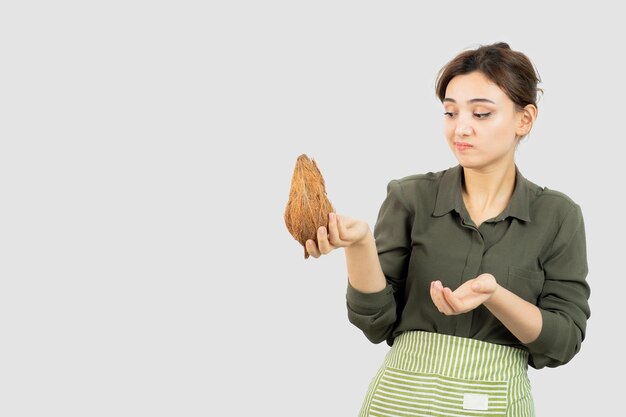 Retrato de mujer joven en delantal sosteniendo un coco contra la pared blanca