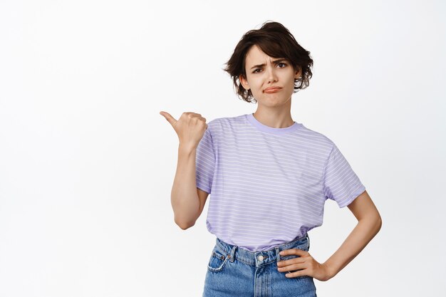 Retrato de mujer joven disgustada, cliente frunciendo el ceño apuntando a la izquierda con expresión de cara escéptica, disgusto, opinión negativa, mala retroalimentación, de pie en blanco.