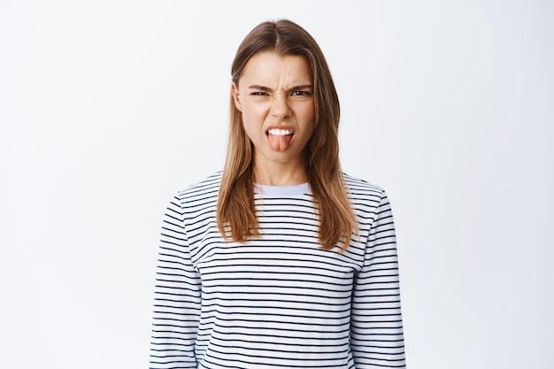 Retrato de mujer joven disgustada expresa aversión y desdén, mostrando la lengua y entrecerrando los ojos por la aversión a algo malo, de pie en blanco