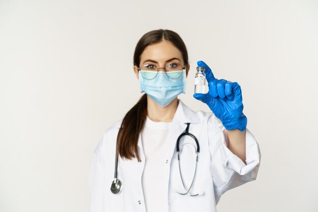 Retrato de mujer joven, doctora con mascarilla médica y uniforme, mostrando vacuna, campaña de vacunación covid-19, de pie sobre fondo blanco.