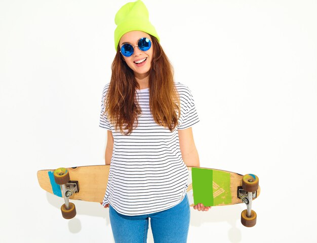 Retrato de mujer joven elegante modelo en ropa casual de verano en gorro amarillo posando con escritorio de longboard. Aislado en blanco