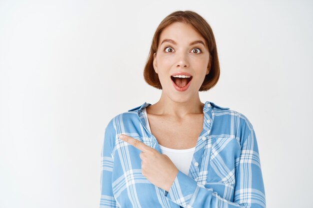 Retrato de mujer joven emocionada apuntando a la izquierda jadeando y diciendo wow asombrado, mira gritando de alegría, de pie en la pared blanca