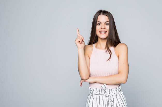 Retrato de mujer joven emocionada de pie señaló con el dedo hacia arriba aislado sobre pared gris