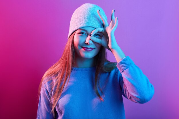 Retrato de mujer joven emocionada vestida de suéter casual y gorro de punto, mostrando el signo de ok contra su ojo