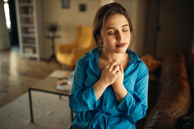 Foto gratuita retrato de mujer joven encantadora alegre sentada en el sofá en la sala de estar cerrando los ojos, manteniendo las manos en el pecho, rezando, con expresión facial tranquila y pacífica, soñando con algo agradable