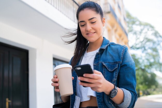 Retrato de mujer joven escribiendo en el teléfono y sosteniendo una taza de café mientras está de pie al aire libre en la calle