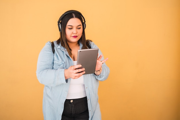 Retrato de mujer joven escuchando música con auriculares y tableta digital al aire libre contra la pared amarilla