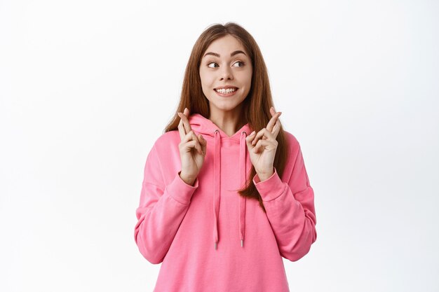 Retrato de mujer joven esperanzada hace su deseo, cruza los dedos para la buena suerte, sonríe y mira a un lado el texto promocional, se encuentra contra la pared blanca