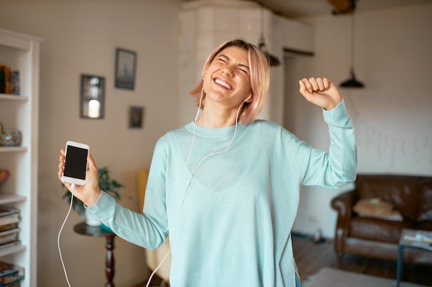 Retrato de mujer joven con estilo divertido con cabello rosado posando en el interior del acogedor apartamento en auriculares