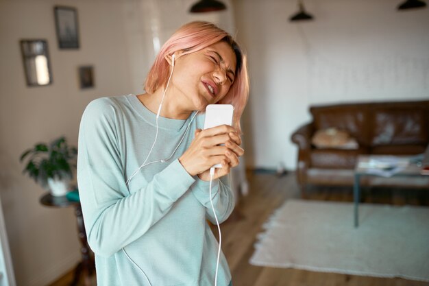 Retrato de mujer joven con estilo divertido con cabello rosado posando en el interior del acogedor apartamento en auriculares