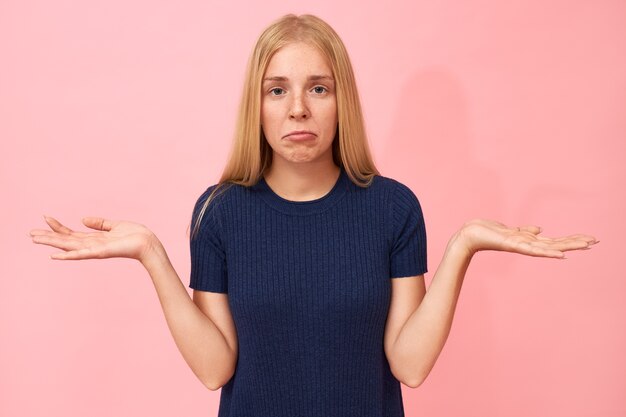 Retrato de mujer joven con estilo con el pelo rubio lacio haciendo pucheros, encogiéndose de hombros, manteniendo los brazos extendidos, sintiéndose decepcionada por los planes arruinados, sintiéndose perdido