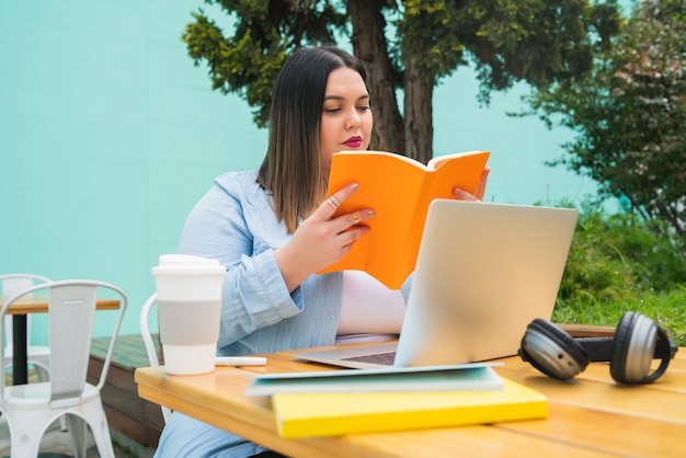 Retrato, de, mujer joven, estudiar, con, computadora portátil, y, libros, mientras, sentado, aire libre, en, cafetería
